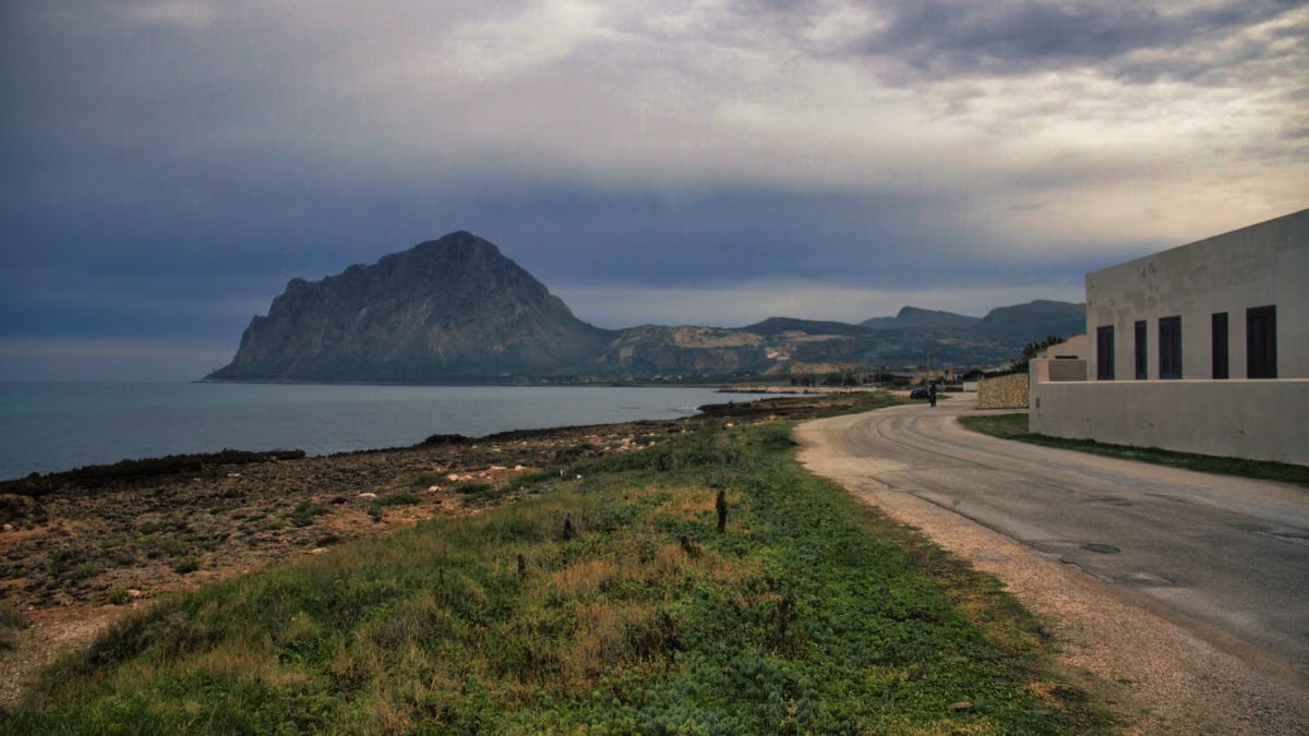 Tranquil coastal road with Monte Cofano in Sicily, Italy under cloudy skies.